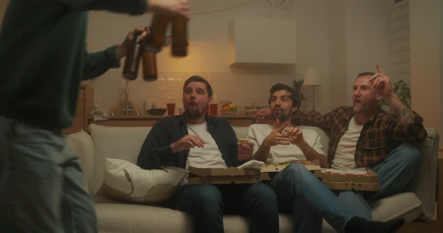 A cheerful guy brings bottles of beer to his friends who are sitting on the sofa during a cheerful bachelor party at home in the evening in the kitchen