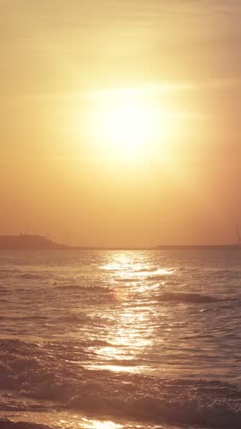 Orange sun over the beach. Sunset on a calm sea. Silhouette of a seaport in the distance. Path of light on the waves.