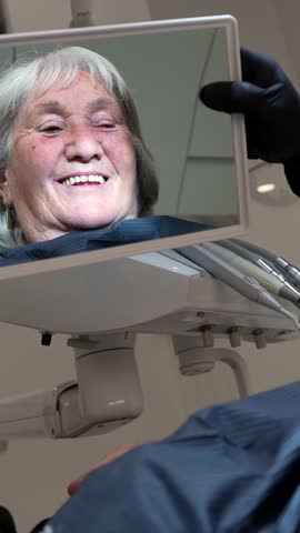 Senior woman checking her teeth in the mirror at the dental clinic, happy with the results of the treatment