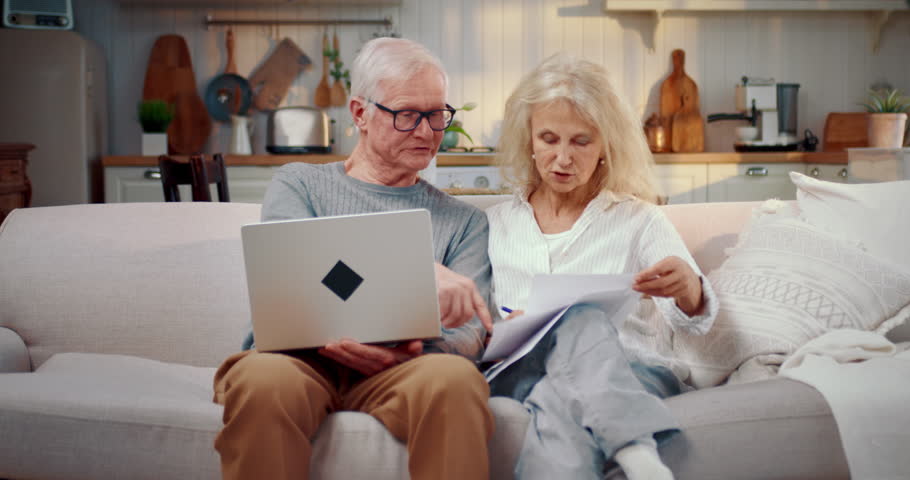 Elderly spouses using internet for pay utility bills online, portrait at home. Mature woman holding monthly invoice and discussing with husband, mortgage payment and insurance premiums, portrait shot