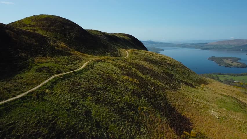 Aerial view above winding mountain road during a beautiful sunny day and blue sky, rural scenery. Majestic rolling hillside, blue freshwater lake with a green island is behind, Scottish loch landscape