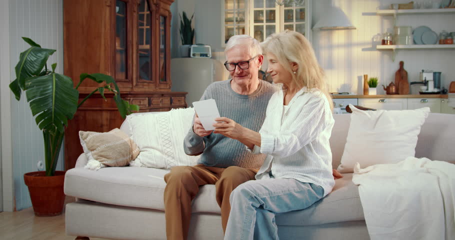 Happy elderly spouses laughing when watching old photos from family archive. Cheerful lady and her husband sitting on cozy couch in living room, interior of house, old married couple have fun together
