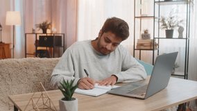 Young Indian man sitting at table joins online class through laptop, attentively listening to lecture, taking notes in notebook. Hispanic guy at home, focused on own task or learning activity nearby. - Powered by Shutterstock - Get 15% off with code: PIKWIZARD15