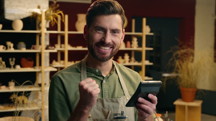 Happy successful Caucasian small business owner man male guy businessman salesman potter holding calculator calculating finance budget success growth money clench fist gesture win luck indoor workshop