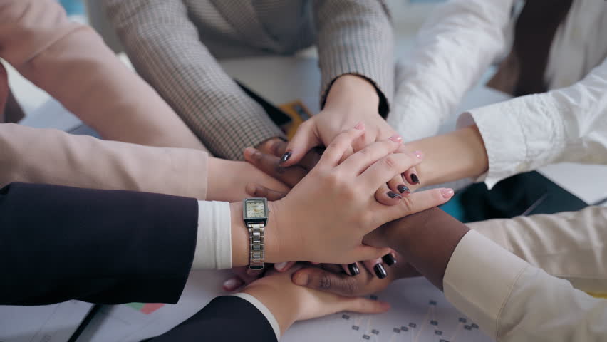 African American Japanese Hispanic multiethnic business women females team girls coworkers businesswomen hands arms folded in unity gesture cooperation teamwork partnership documents table at office