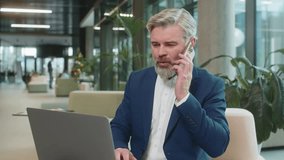 Confident mature businessman in suit answering to client on mobile phone call, using smartphone and laptop. Conversation. Caucasian bearded man guy working online job, talking in modern office lobby. - Powered by Shutterstock - Get 15% off with code: PIKWIZARD15