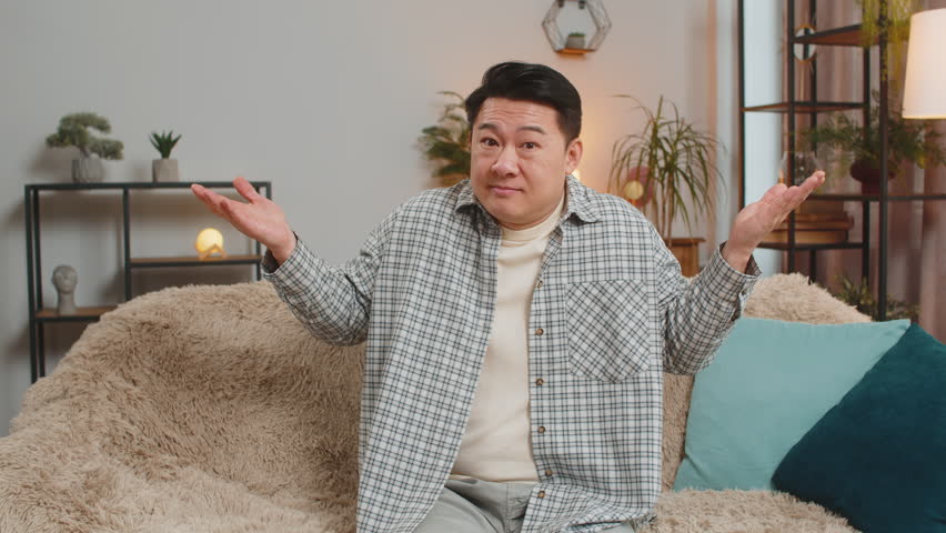 I dont know what to say. Confused Asian handsome man feeling embarrassed about ambiguous question, having doubts, no idea, being clueless and uncertain at home. Chinese guy sits on sofa in living room