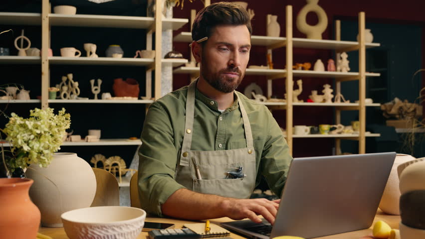 Serious Caucasian skilled man guy small business owner businessman crock potter ceramist artisan male working typing laptop networking computer online shopping ordering material inside workshop studio