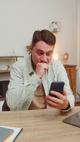 Happy young Caucasian man sitting at table using mobile phone smiling at home office. Guy freelancer texting share messages on smartphone social media applications online watching movie. Vertical.