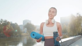 Young happy sportswoman with mat in her hands is heading to training outdoors in park. Smiling optimistic fit woman girl in activewear with headphones and water bottle goes to gymnastics training. - Powered by Shutterstock - Get 15% off with code: PIKWIZARD15
