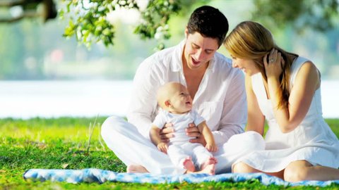 Proud young Caucasian parents smiling at laughing baby son sitting outdoors together on the grass shot on RED EPIC