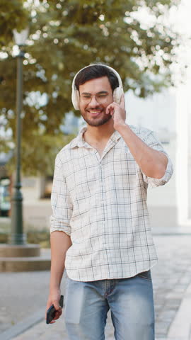 Happy overjoyed young man tourist in wireless headphones choosing, listening favorite energetic disco music on smartphone outdoors. Indian tourist guy walking passes by urban sunny city town street.