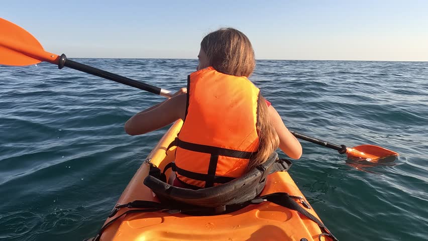 A young girl in an orange kayak paddles across the rippling blue water, their hand reaching for the paddle. The vast ocean stretches out in the distance. Summer holiday vacation and travel concept