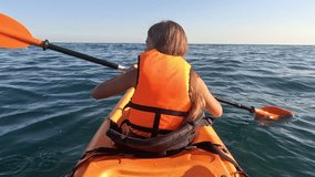 A young girl in an orange kayak paddles across the rippling blue water, their hand reaching for the paddle. The vast ocean stretches out in the distance. Summer holiday vacation and travel concept - Powered by Shutterstock - Get 15% off with code: PIKWIZARD15