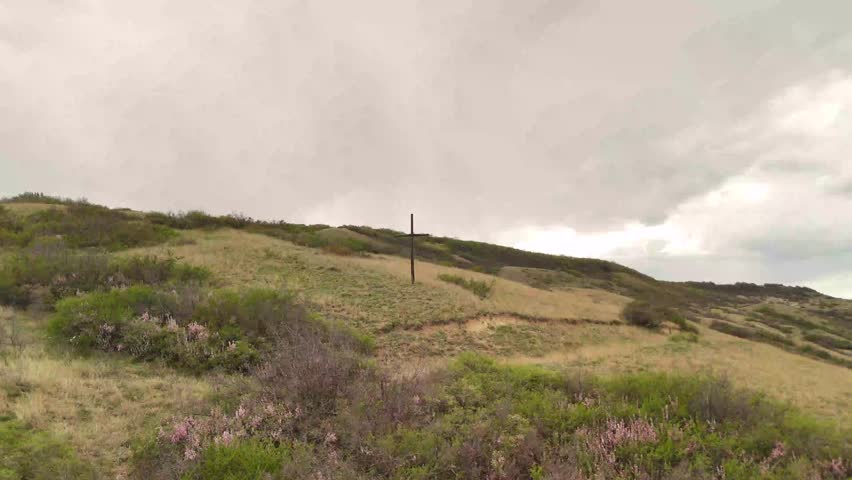 Weathered wooden cross rising from verdant grassy slopes, cloudy sky casting somber shadows over serene pastoral landscape evoking quiet spiritual reflection