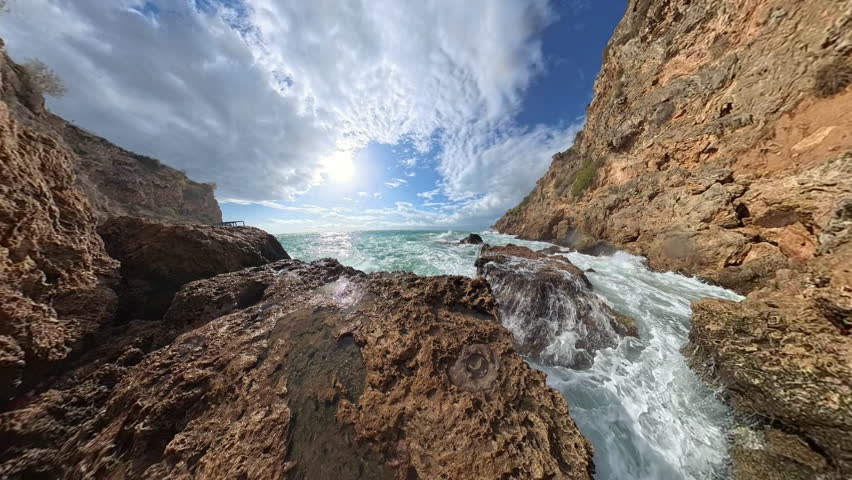 A stunning wide-angle capture of powerful waves breaking into the open sea, with sunlight glinting off the water under a brilliant blue sky dotted with clouds