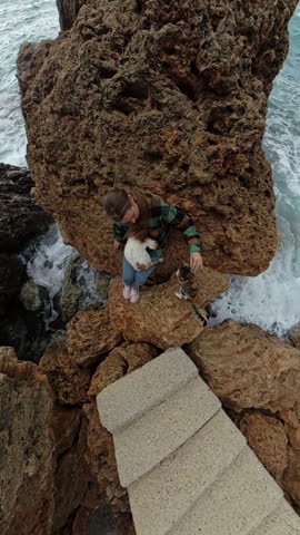 Vertical video. A woman on jagged rocks near the sea interacting with two cats, with rough coastal textures and ocean waves in the background