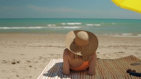 Young woman in swimsuit and sunhat lounging on striped beach towel, basking under sunlight with ocean waves rolling softly in background - Powered by Shutterstock - Get 15% off with code: PIKWIZARD15