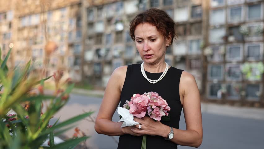 Sorrowful young woman in black dress with bouquet of flowers and handkerchief in hands, stands in cemetery. Visiting grave of deceased relative, mourning dead loved one. Appeal to God, prayer for dead