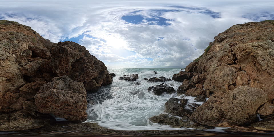 Immersive 360-degree view of crashing waves in a narrow rocky cove, surrounded by jagged cliffs and a bright, partly cloudy sky