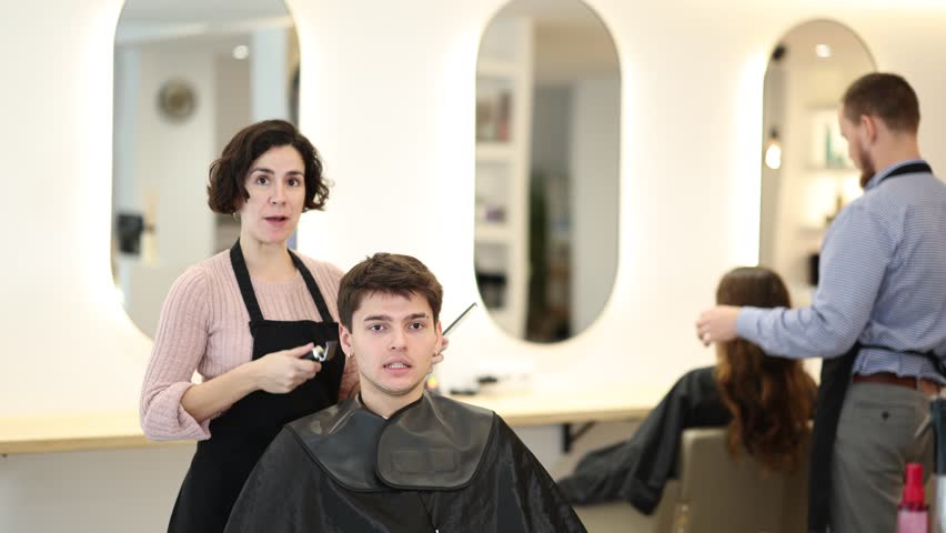 Adult woman hairdresser stylist gives haircut to young guy client in beauty salon
