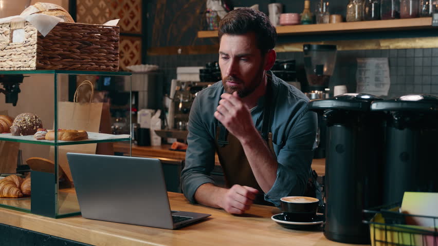 Pensive Caucasian entrepreneur barista employee male browsing Internet app laptop commerce small business owner thinking in cafeteria manager choosing guy online shopping cafe man waiter typing gadget