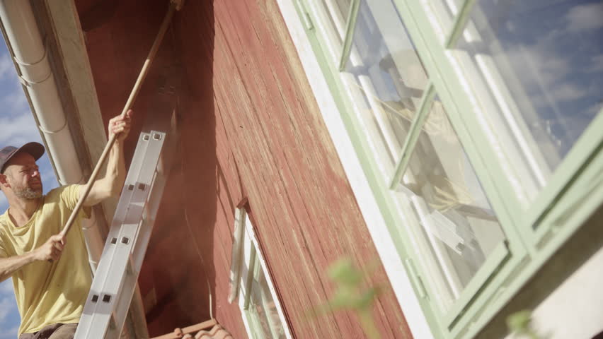Man on ladder against wooden house uses broom to brush off peeling red paint