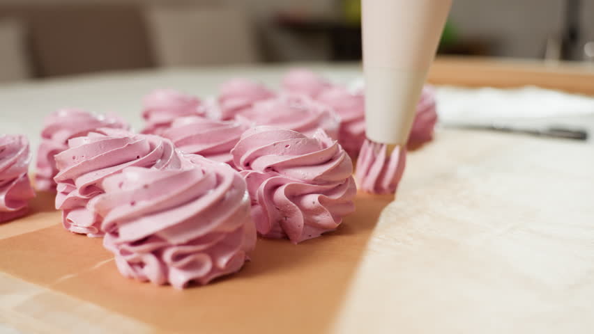 Close up of cupcake preparation process showing piping bag squeezing swirls of pink dough onto brown baking paper, cupcakes aligned in neat row ready for baking, soft focus background