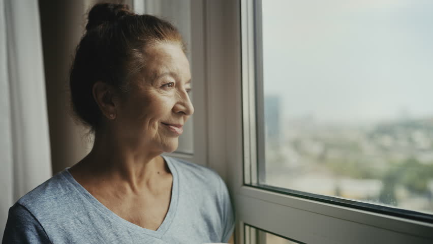 Relaxed senior woman drinking coffee and looking out the window, in a good mood