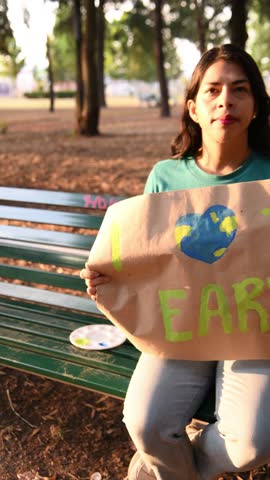 Hispanic Latina brunette woman holding an earth day sign on a bench in urban park. Message of ecology and sustainability at sunset.