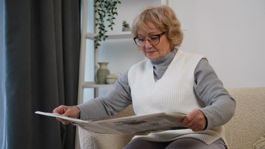 Serious elderly woman reading newspaper at home