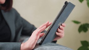 Businesswoman in grey suit using digital tablet on couch with hand gesture mid-air, possibly interacting with screen or video calling, indoors in relaxed work-from-home or casual business setting - Powered by Shutterstock - Get 15% off with code: PIKWIZARD15