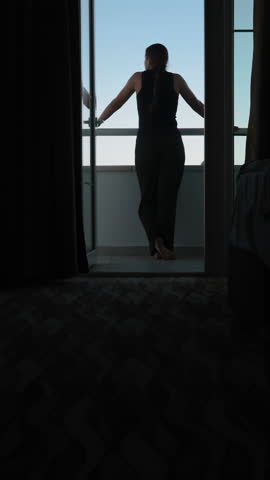 Vertical video. A dark bedroom with light from the blue sky coming through the open balcony door where a woman stands and enjoys the sea views.