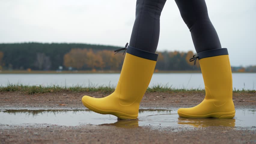 Woman wearing bright yellow rubber boots walking along the path splashing through puddles near serene lake during overcast autumn overcast day. Experiencing carefree freedom of outdoor exploration