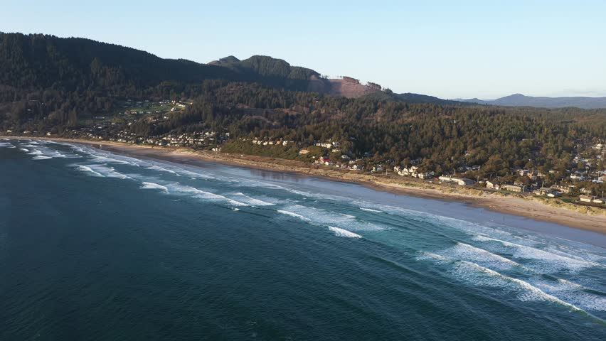 4K aerial drone video of Manzanita Beach on the Oregon Coast, showing waves of the Pacific Ocean, small coastal town, sunset, sandy shoreline, forested mountains. Perfect for travel and tourism themes