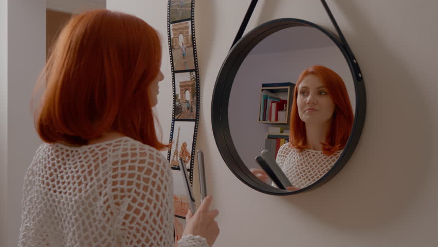 Young woman with vibrant red hair straightens her locks with a flat iron while enjoying her morning routine in the comfort of her home. She focuses on beauty and self-care.