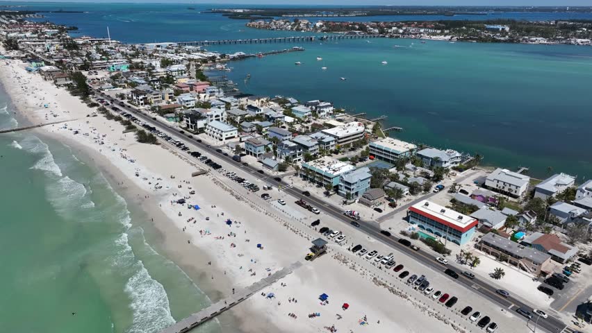 Jetty and people enjoying sunlight at sandy beach of Bradenton. Clear water with reaching waves. Luxury homes and buildings in small town of Florida, USA. Aerial tilt up wide shot.