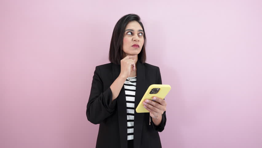 A thoughtful pondering South Asian young woman, looking at her phone with disappointed serious face expression, isolated over pink background