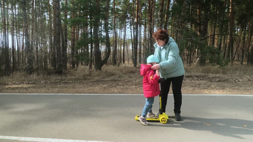 Little girl riding scooter while walking with her grandmother in the forest on a sunny day
