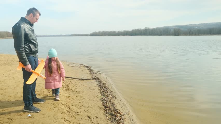 Dad and daughter walking near lake share holding airplane toy on sunny day