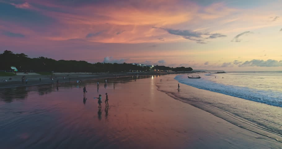 Tourists walking on Dreamland Beach, Bali, Indonesia, during a colorful sunset, with a traditional fishing boat gently rocking on the waves, creating a serene and picturesque scene