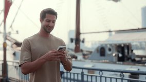Portrait of smiling Latin American man texts on smartphone standing against luxury yacht. Hispanic guy writes message on cellphone walking in city port. Device for communication - Powered by Shutterstock - Get 15% off with code: PIKWIZARD15