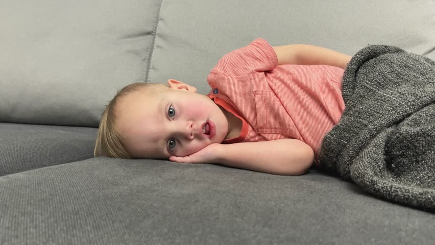 A young child lounging on a sofa, looking cozy with a gray blanket, capturing a moment of relaxation.