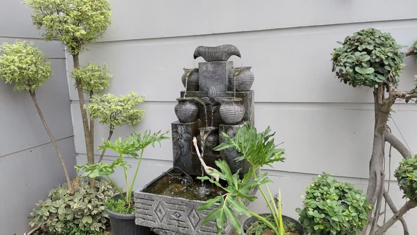 A small tiered fountain with urns, surrounded by potted plants against a grey wall