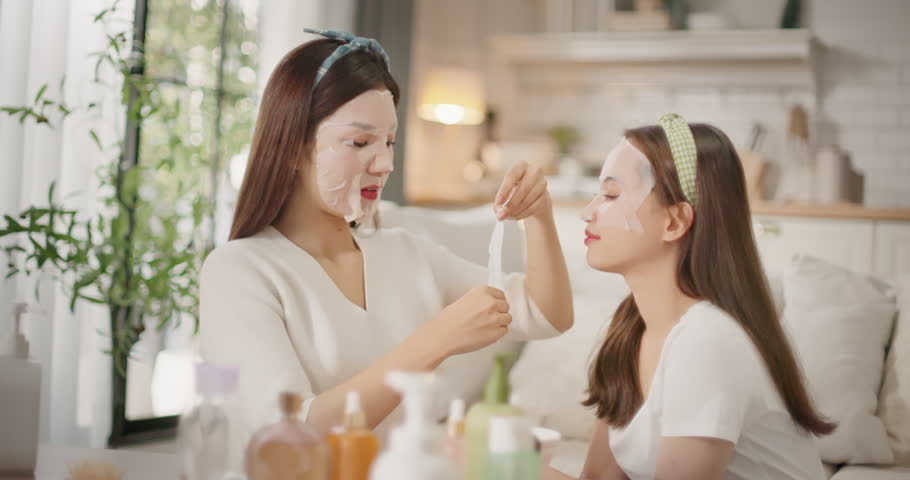 Two Asian women friends applying sheet masks together during beauty daily routine at home. Girl talk, skincare bonding moment, Gen Z lifestyle, self care and relaxing feminine friendship vibe.