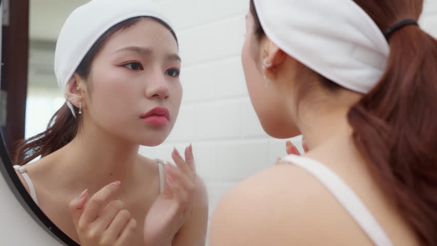Beautiful young asian woman examining skin of facial while looking mirror in bathroom at home, unhappy woman touching cheek with check wrinkle with stress and worry, beauty and skincare concept.