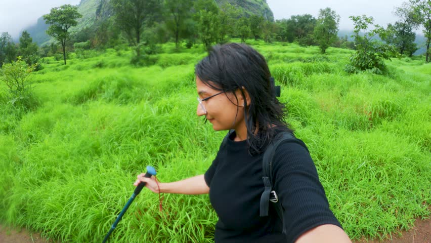 4k Selfie video of Indian young woman with backpack hiking in green forest of Sahyadri hills in Maharashtra, India. Monsoon trekking, active lifestyle concept. Girl hiking outdoors holding hiking pole
