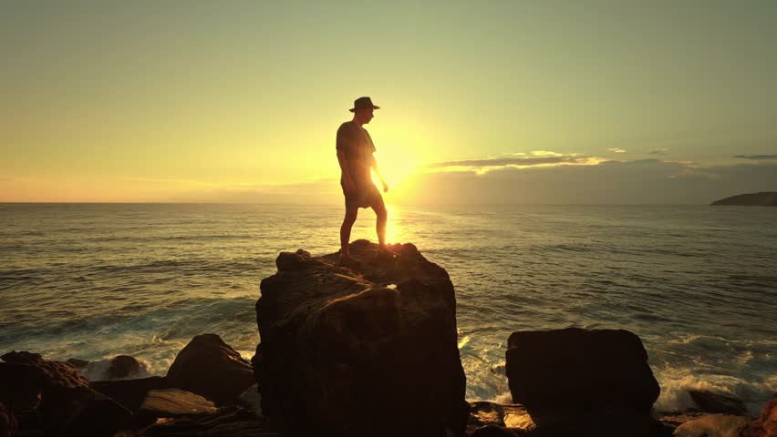The silhouette of a man in a hat stands atop a large oceanfront rock against a breathtaking sunrise sunset sky. This inspiring seascape captures the magical transition between night and day