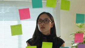 Young businesswoman wearing eyeglasses is looking at colorful sticky notes attached to a glass wall in the office, brainstorming new ideas for her business - Powered by Shutterstock - Get 15% off with code: PIKWIZARD15