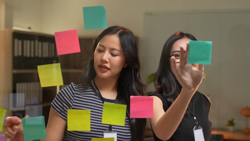 Two young asian businesswomen are using colorful sticky notes on a glass wall to brainstorm and plan their business strategy in a modern office